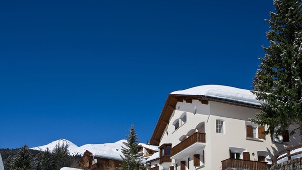 Un edificio imbiancato dalla neve con numerosi balconi, circondato da alberi e con una montagna sullo sfondo.