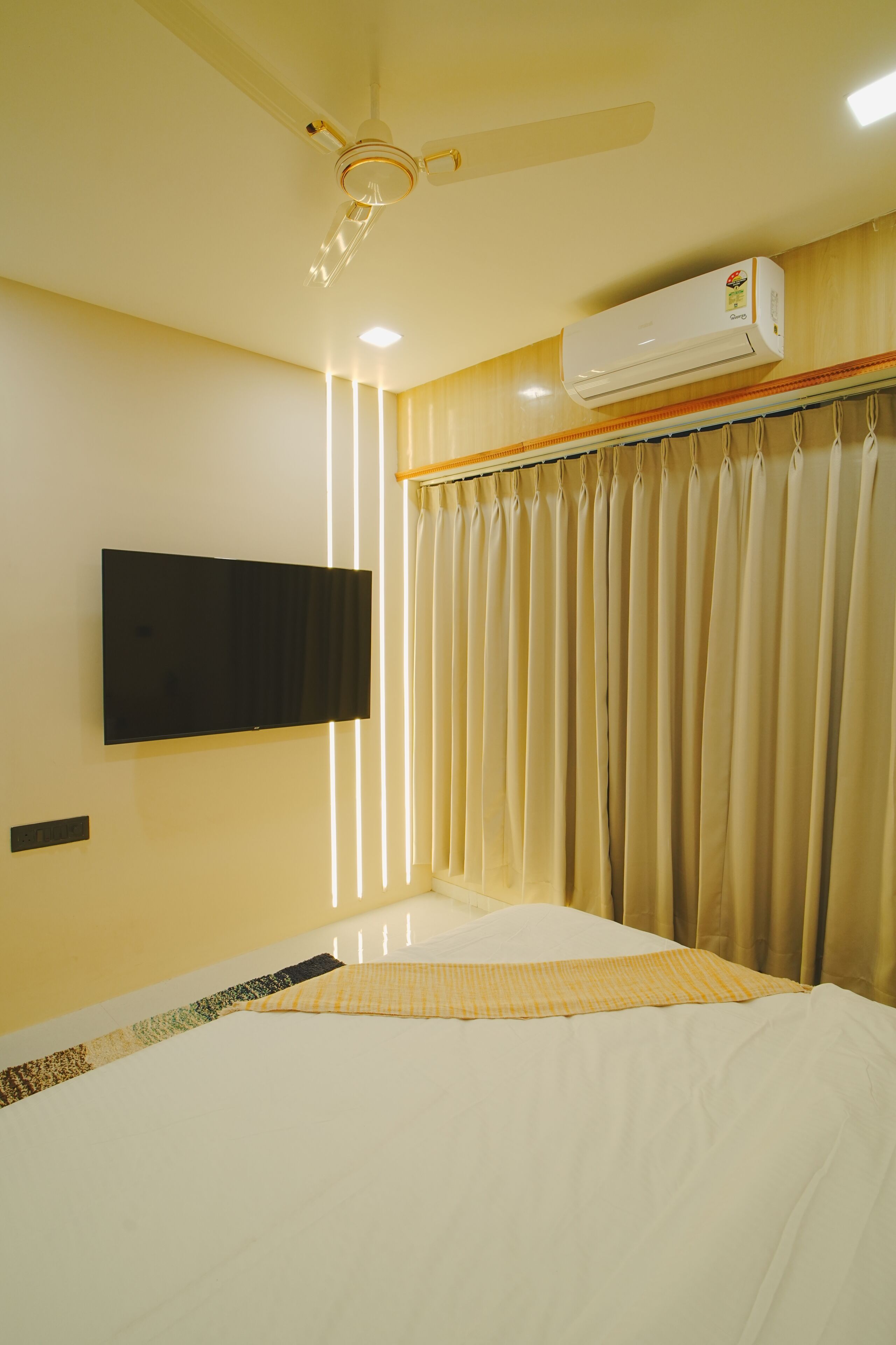 A hotel room with a bed, a flat-screen TV mounted on the wall, an air conditioning unit, and a ceiling fan.