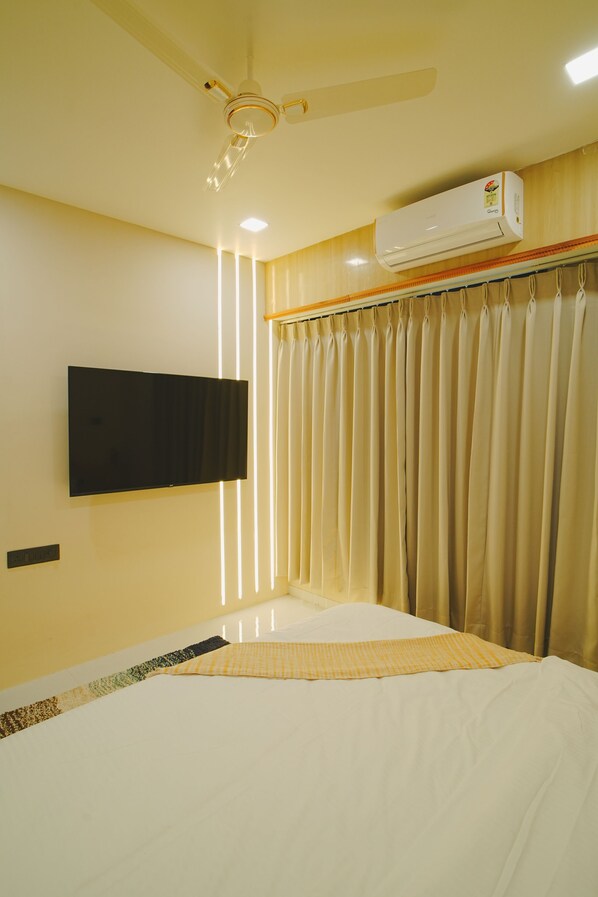A hotel room with a bed, a flat-screen TV mounted on the wall, an air conditioning unit, and a ceiling fan.