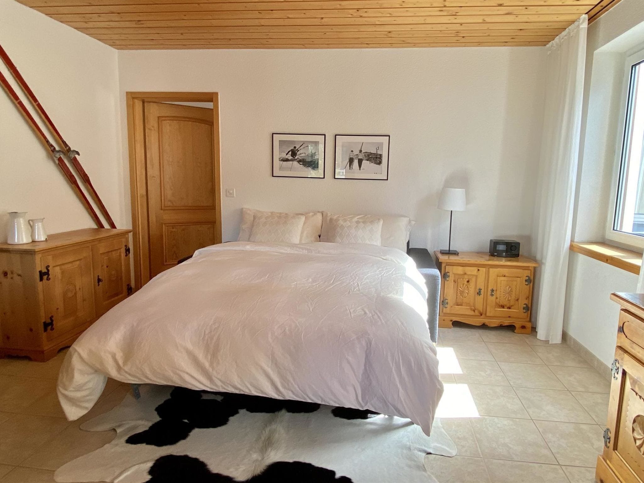 A bedroom with a large bed, wooden wardrobe, a nightstand with a lamp, and two framed pictures on the wall.