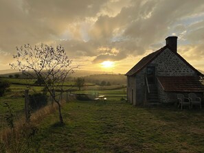 A rustic stone building with a sloped roof, a wooden fence, and a tree in the foreground.