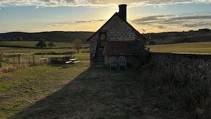 A stone cottage with a chimney, a small staircase, and two chairs outside.