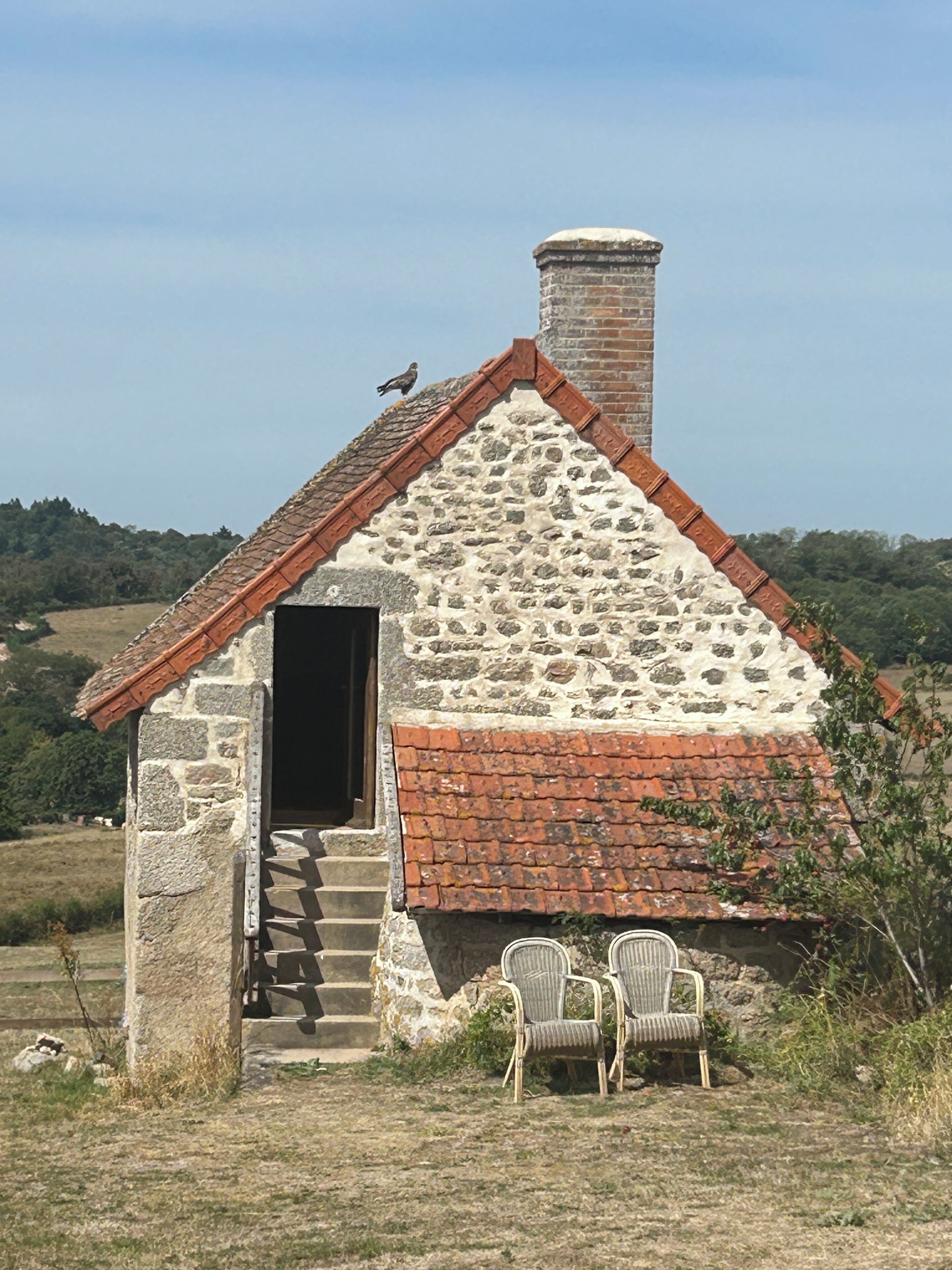 Una cabaña de piedra con chimenea, una puerta pequeña y dos sillas de mimbre afuera.