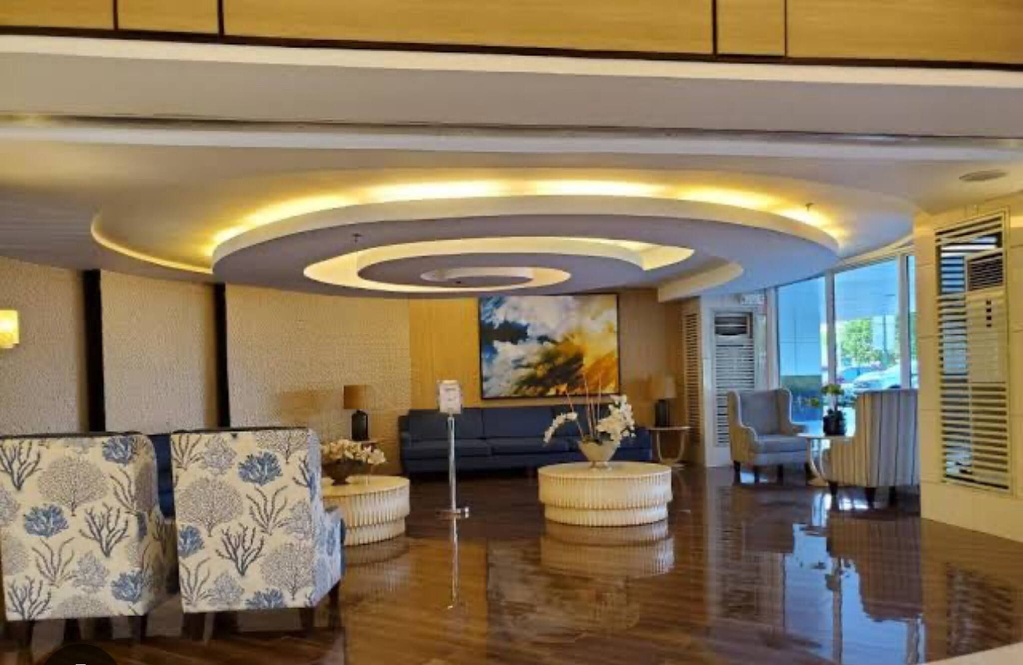 Un vestíbulo de hotel con diseño moderno, que presenta una zona de estar central con un sofá y pufs, un cuadro abstracto grande en la pared, y un ventanal con vistas al exterior.