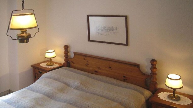 A bedroom with a wooden headboard, a single bed with a striped blanket, two bedside tables with lamps, and a framed picture on the wall. - Casa Laghetto 5 Appartement/Fewo, Bad, WC, 4-Bettwohnung 4-Bettwohnung by Interhome (Arosa)