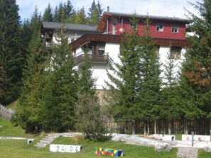 A multi-story building with a red facade, surrounded by trees and a grassy area with a small playground. - Casa Laghetto 5 Appartement/Fewo, Bad, WC, 4-Bettwohnung 4-Bettwohnung by Interhome (Arosa)