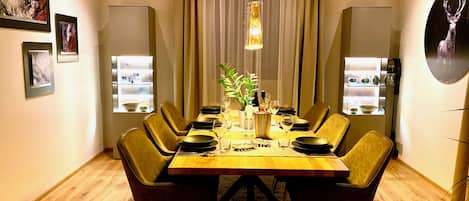 A dining area with a wooden table set for six, surrounded by chairs, with a plant centerpiece and a bottle of wine.