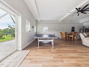 Un vaste espace de vie comprenant une table de ping-pong, un plancher en bois, un ensemble de salle à manger et une grande fenêtre donnant sur un paysage verdoyant.