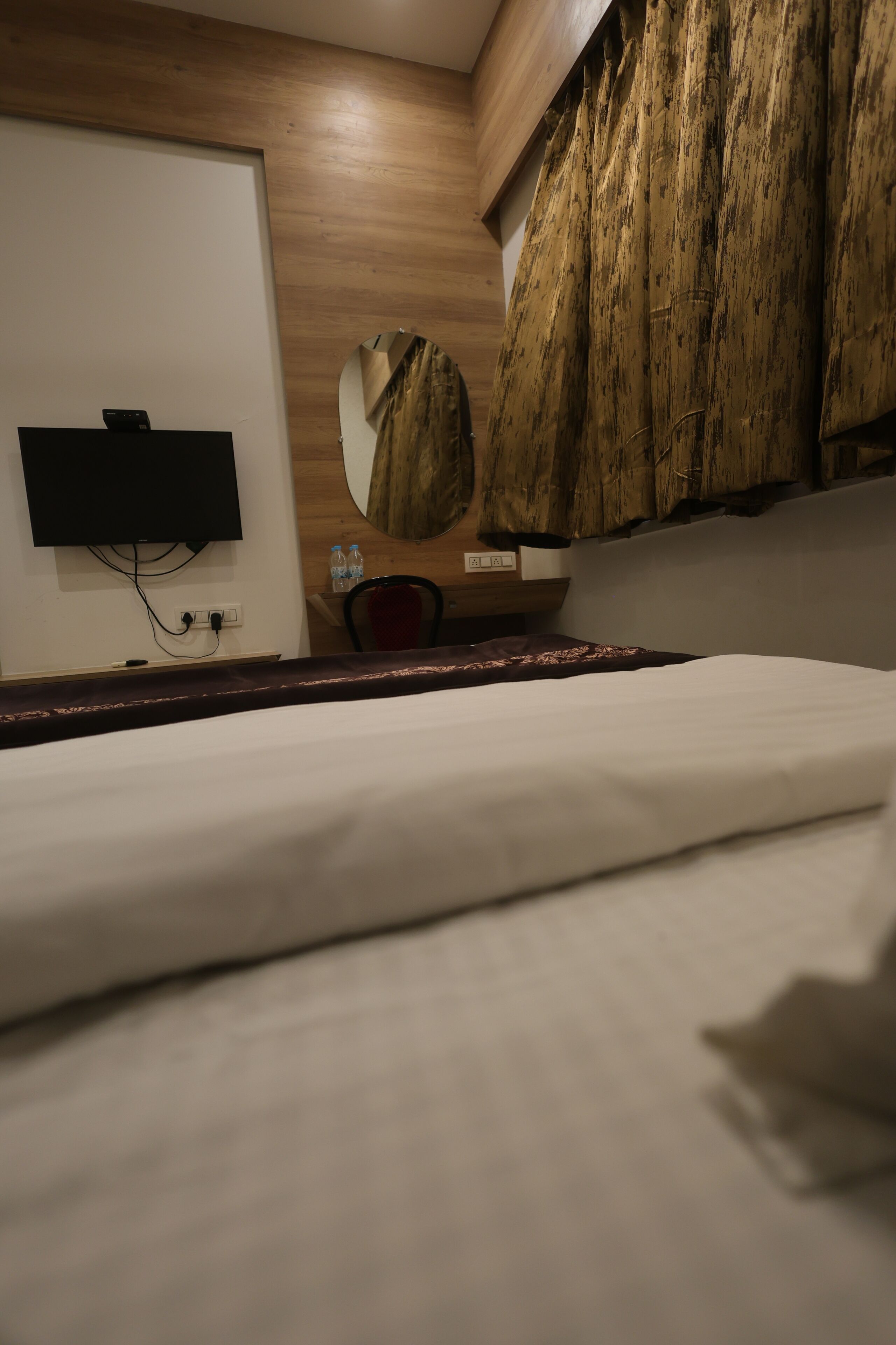 Habitación de hotel con una cama, un televisor de pantalla plana montado en la pared, un armario de madera y un espejo redondo.