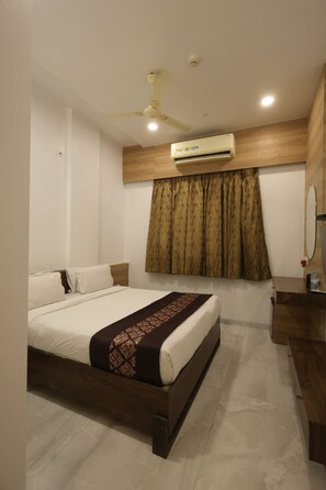Habitación de hotel con cama, ventilador de techo, equipo de aire acondicionado, cabecera de madera, cómoda de madera y espejo.