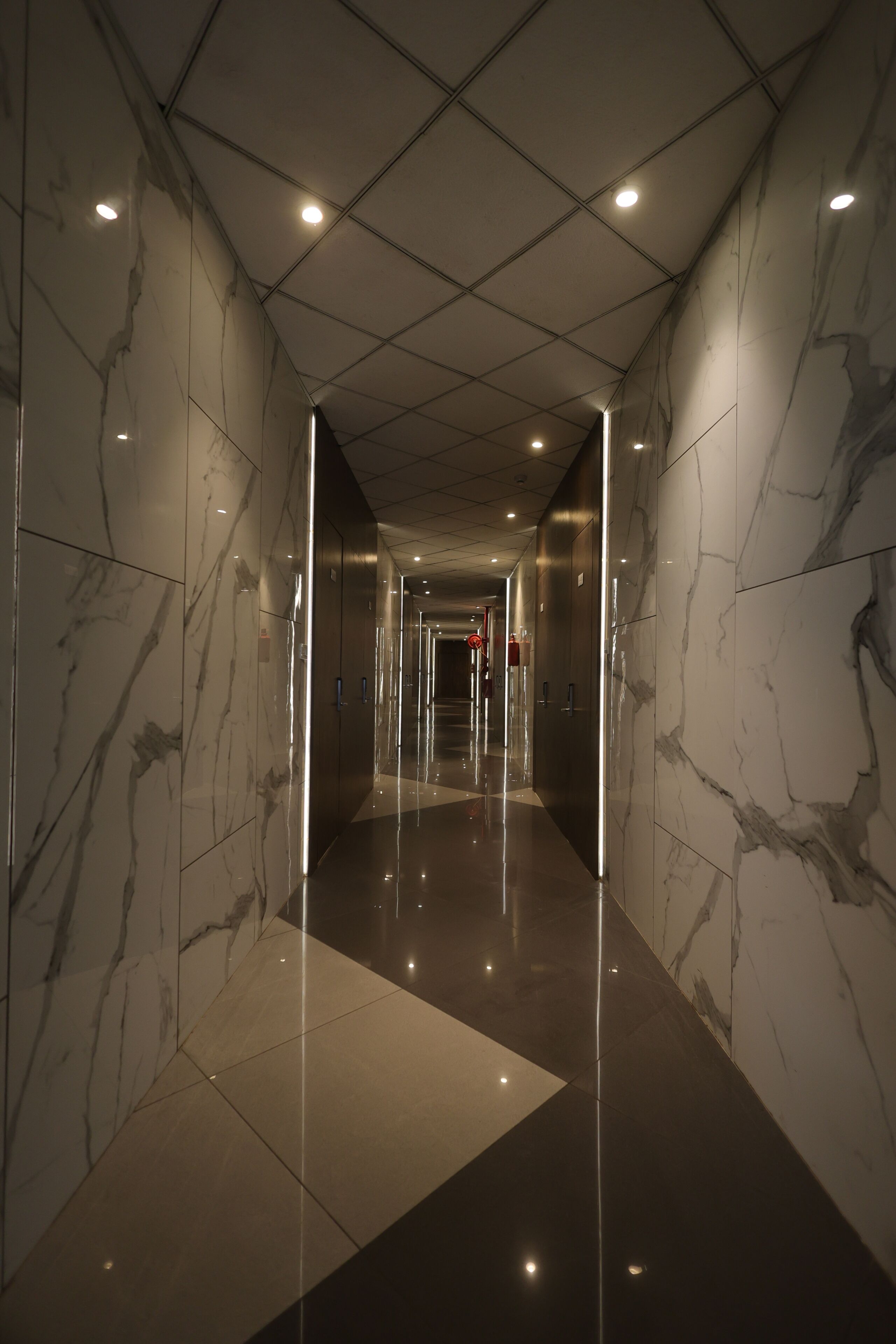 Corredor com paredes de mármore refletoras e chão polido, conduzindo a uma porta com placa de saída de emergência vermelha.