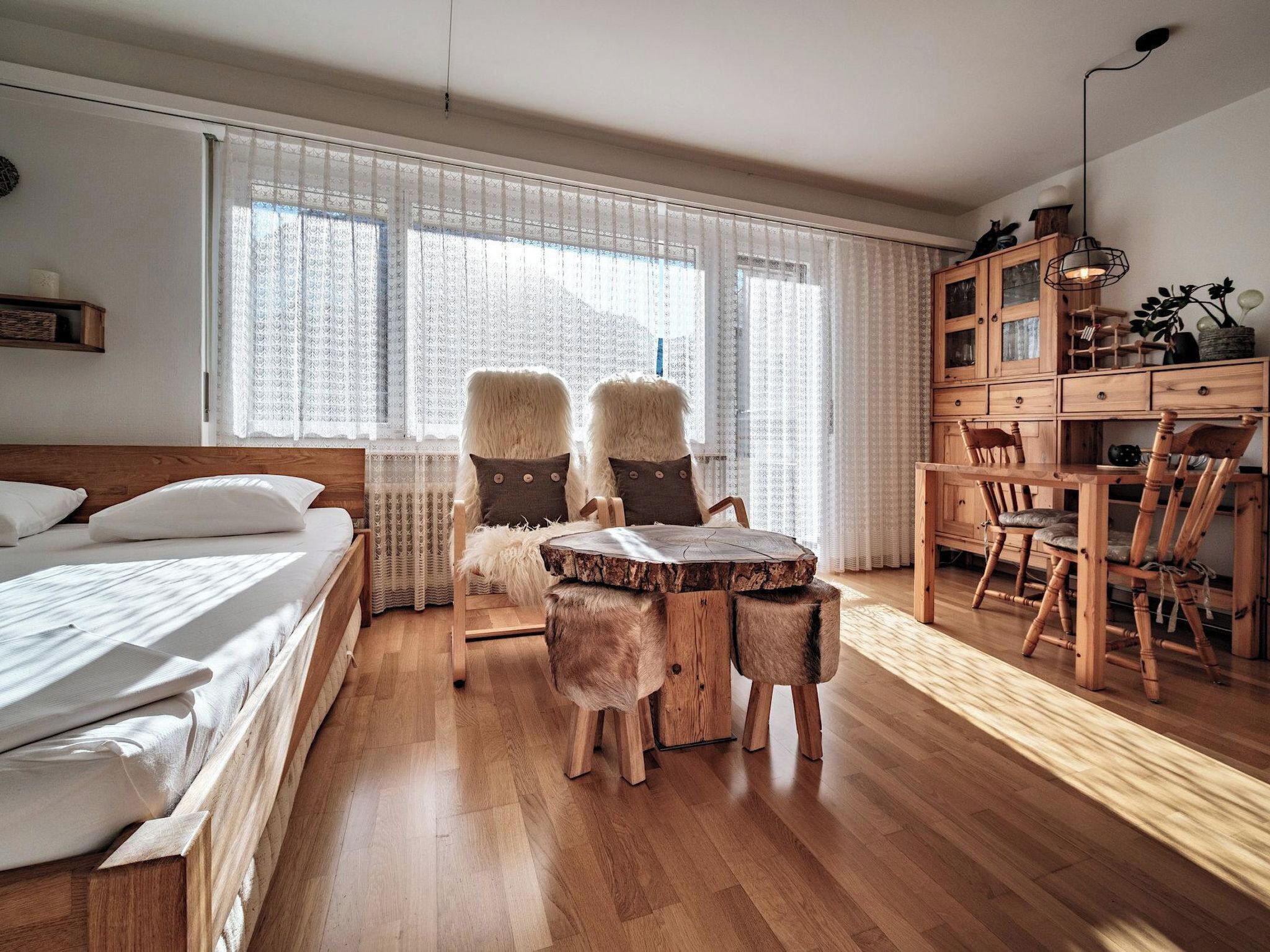 Ein Schlafzimmer mit einem hölzernen Bett, einem runden Holztisch und einem Stuhl. Es gibt eine Holzkommode mit Schubladen und eine Deckenleuchte.