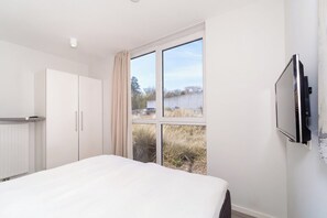 A bedroom with a bed, a wardrobe, a television mounted on the wall, and a window with a view of the outside. - Dünenvilla 10, Apartment 02 (Lübeck-Travemünde)
