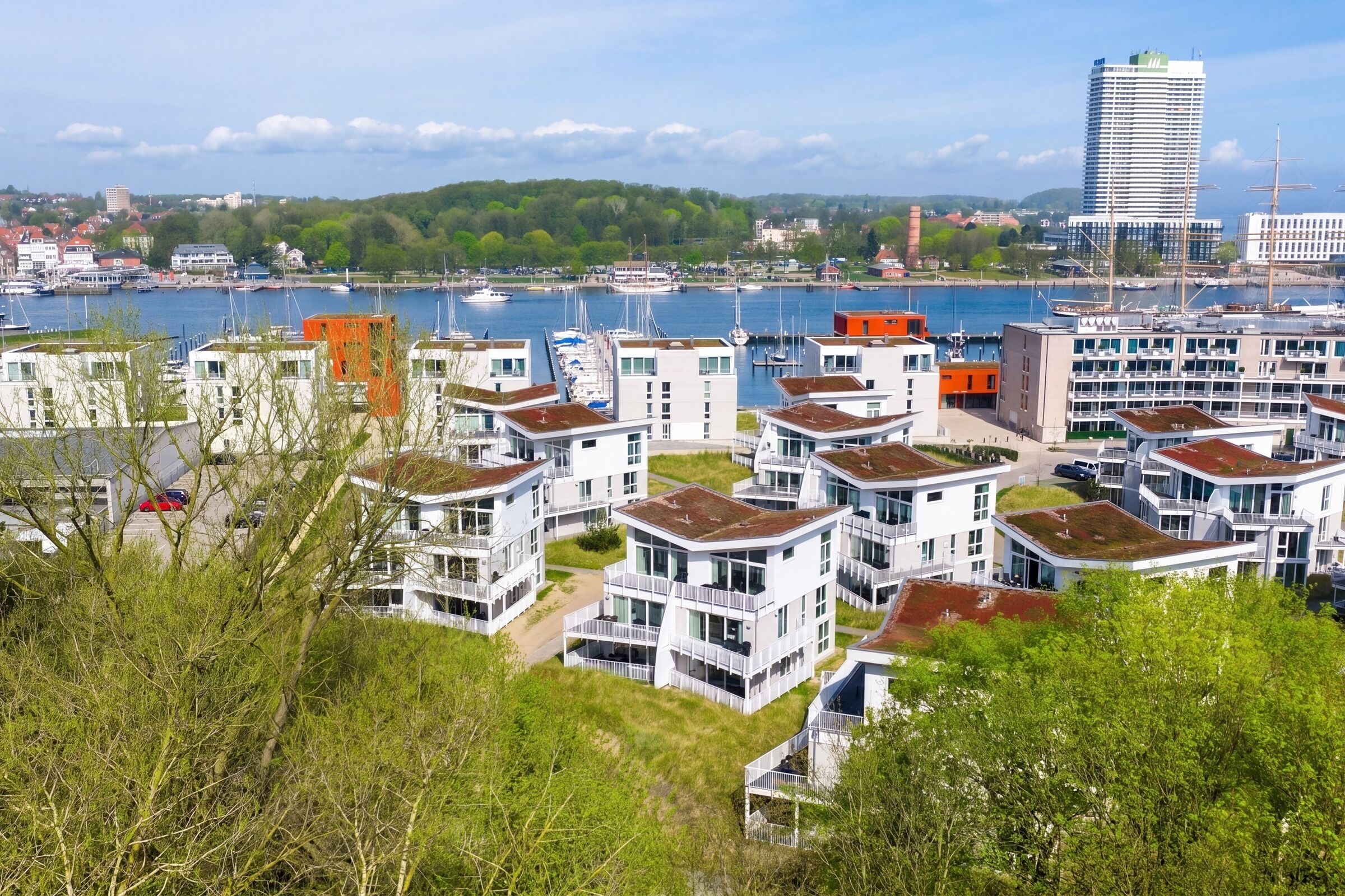 Een kustwoonwijk met moderne witte gebouwen, een jachthaven met boten, en een groene heuvel op de achtergrond.