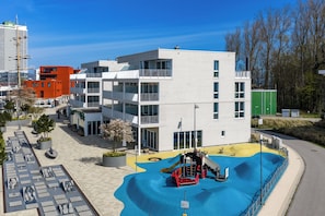 Ein modernes Mehrgeschosshaus mit Spielplatz, Rutsche und Kletternetz sowie einer gepflasterten Fläche mit Bänken.