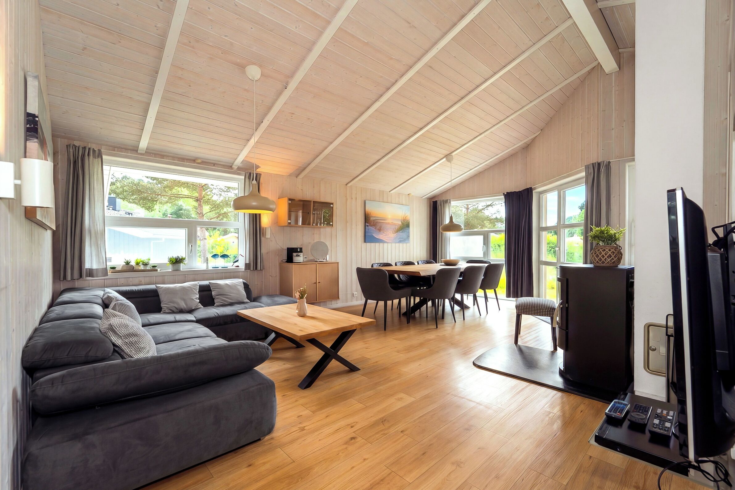 Amplia sala de estar con sofá, mesa de comedor y sillas, piso de madera y ventanas grandes con vistas a la vegetación.