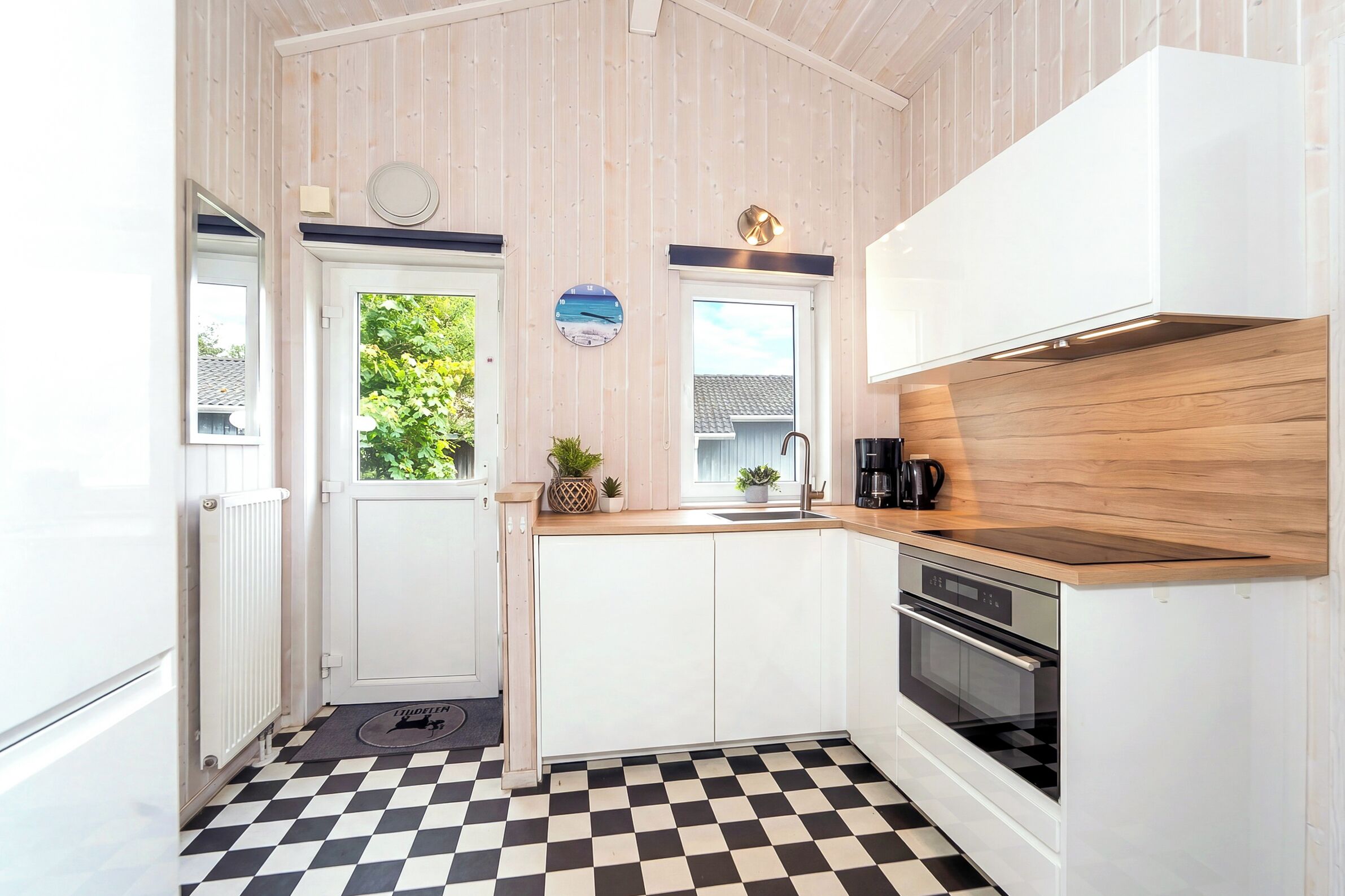 Cocina pequeña con gabinetes blancos, piso a cuadros blanco y negro, encimera de madera y vista a plantas fuera.