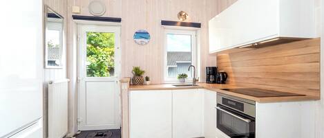 Cocina pequeña con gabinetes blancos, piso a cuadros blanco y negro, encimera de madera y vista a plantas fuera.