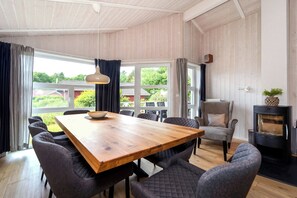 A spacious dining area with a wooden table and chairs, a view of the outdoors through large windows, and a wood-burning stove. - Fresh Breeze Vacation Home 23 (Lübeck-Travemünde)
