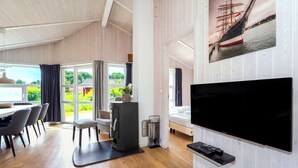 A modern room with a large TV, wooden flooring, a dining area with chairs, and a view of greenery through the windows. - Fresh Breeze Vacation Home 23 (Lübeck-Travemünde)