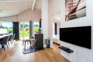 A modern room with a large TV, wooden flooring, a dining area with chairs, and a view of greenery through the windows. - Fresh Breeze Vacation Home 23 (Lübeck-Travemünde)