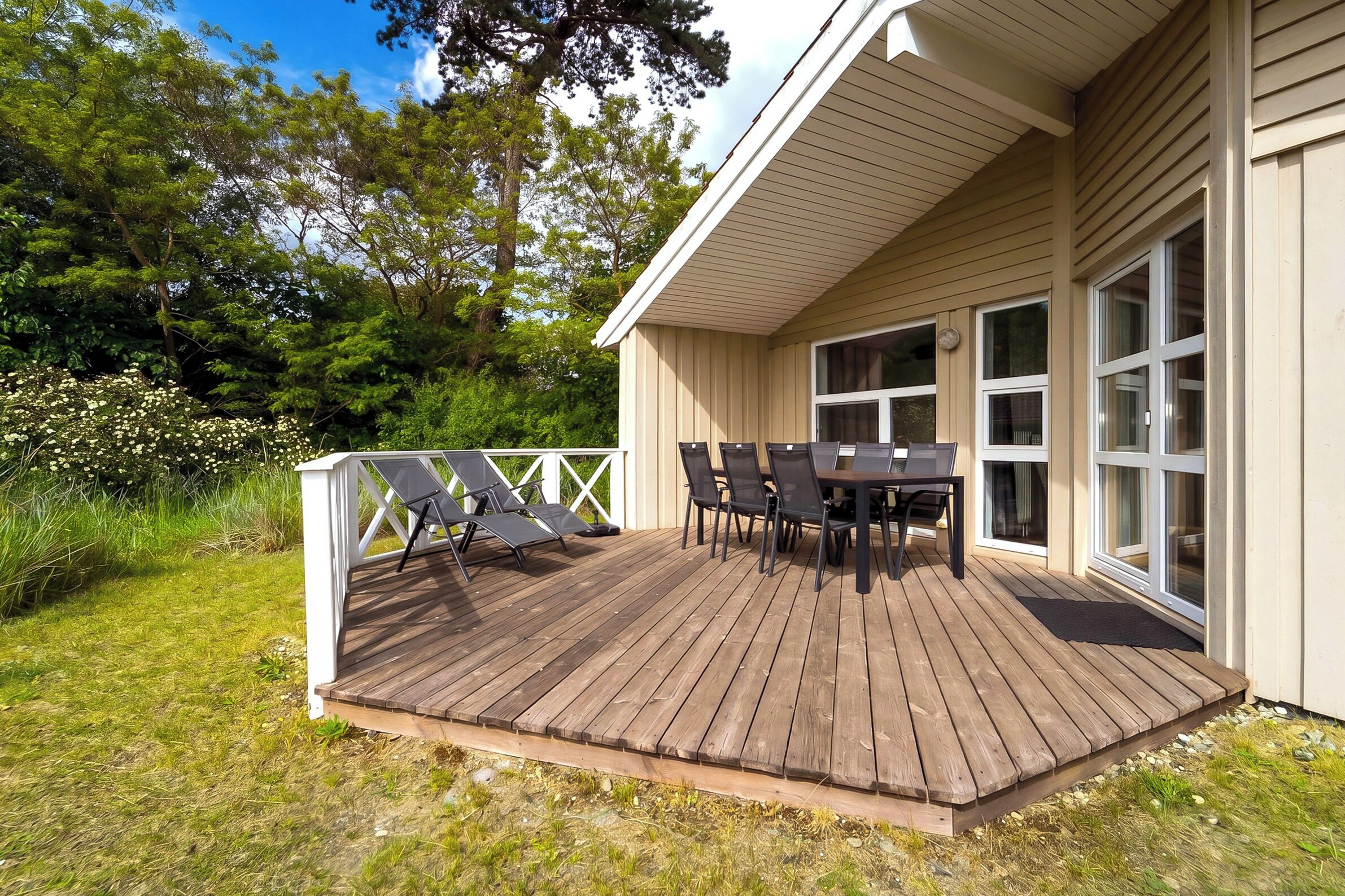 Une terrasse en bois avec des meubles d’extérieur, une balustrade blanche et un bâtiment doté de grandes fenêtres.