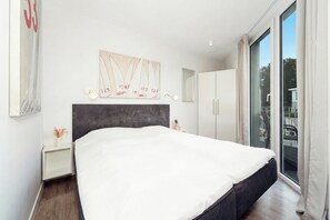 A modern bedroom with a large bed, a headboard, a nightstand, a bedside table, a mirror, and a window with curtains. - Dünenvilla 18, Apartment 05 (Lübeck-Travemünde)