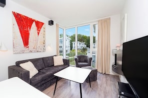 A modern living room with a grey sofa, a white coffee table, a grey armchair, a flat-screen TV, and a large window with curtains. - Dünenvilla 18, Apartment 05 (Lübeck-Travemünde)