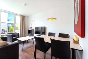 A modern living room with a dining area, a flat-screen TV, and a large window with curtains. - Dünenvilla 18, Apartment 05 (Lübeck-Travemünde)