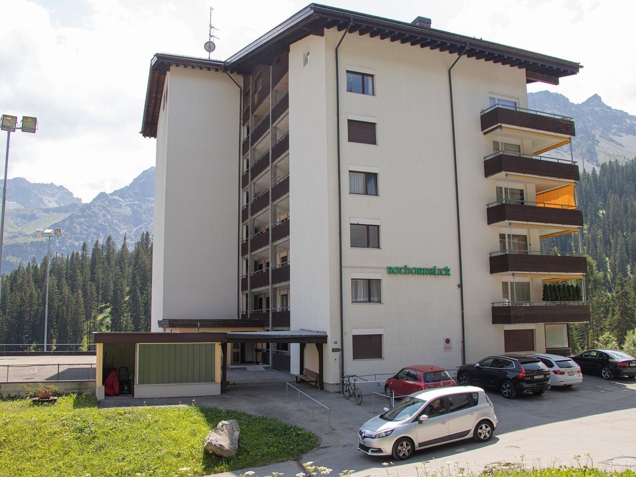 A multi-story building with a sign 'ROCHORNLICK' on the facade, a parking area with cars, and a mountainous backdrop.