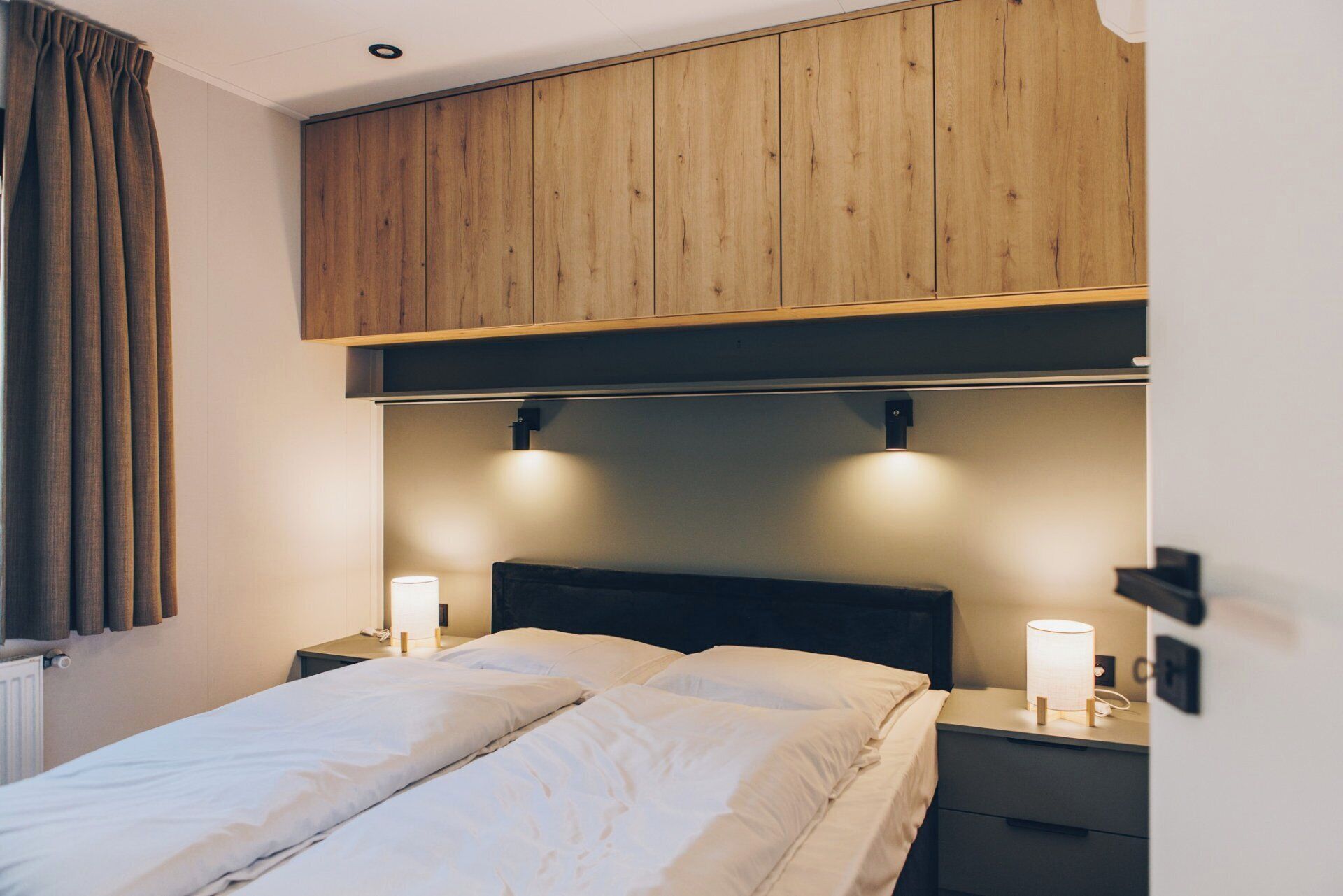 A hotel room with a bed, two bedside lamps, a wooden headboard, and a window with curtains.