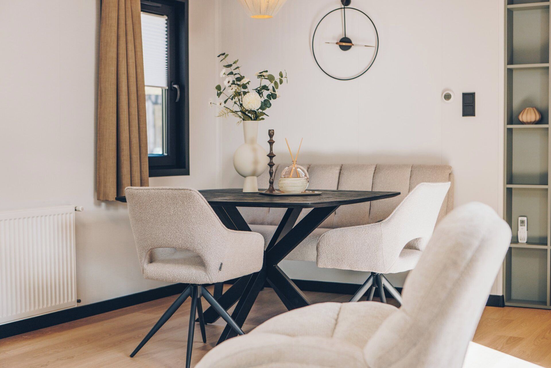 Een moderne eetruimte met een ronde tafel, twee stoelen, een vaas met bloemen en een wandklok.