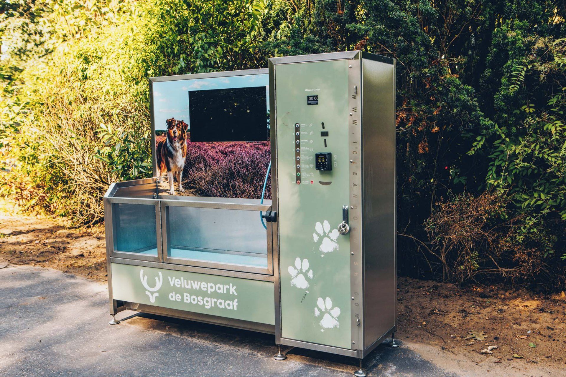 A metal vending machine with a glass window, labeled 'Veluwepark de Bosgraaf,' and a dog standing inside.
