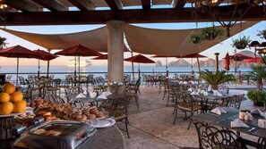 Een terras met tafels en stoelen, een buffet met gebak en fruit, en uitzicht op zee.
