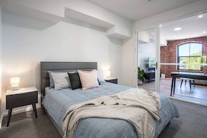 A modern bedroom with a bed, nightstands, and a view of a dining area. - Webb & Co Mid-Rise Apartment (Bendigo)