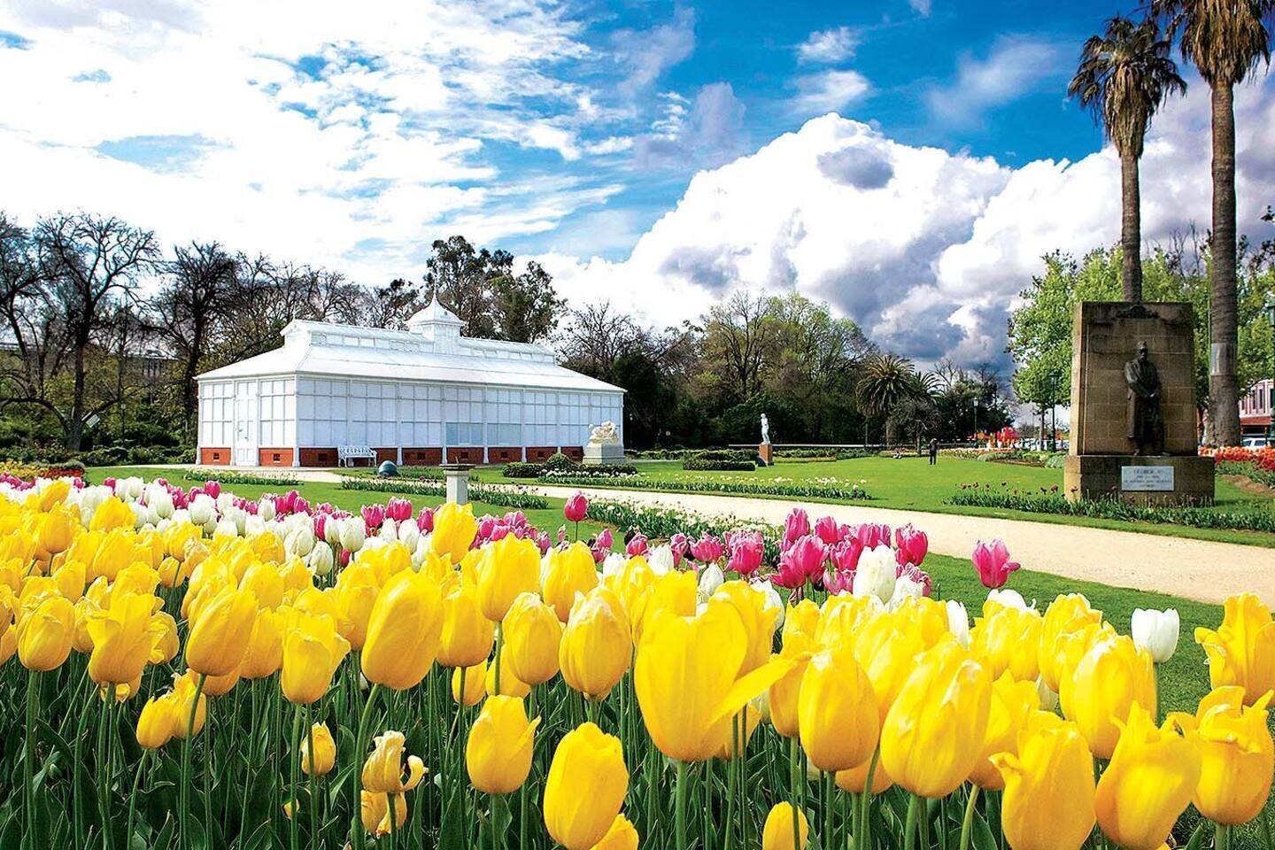 Um edifício com fachada de vidro, rodeado por um jardim com tulipas amarelas e rosas, e uma estátua ao fundo.