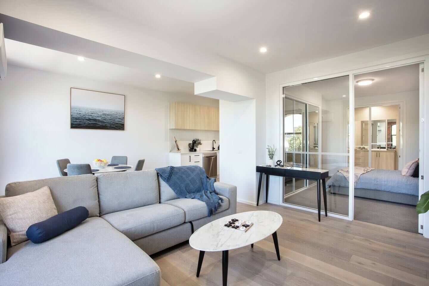 A modern living room with a sofa, coffee table, and a view into the kitchen and bedroom through glass doors.