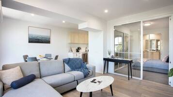 A modern living room with a sofa, coffee table, and a view into the kitchen and bedroom through glass doors.