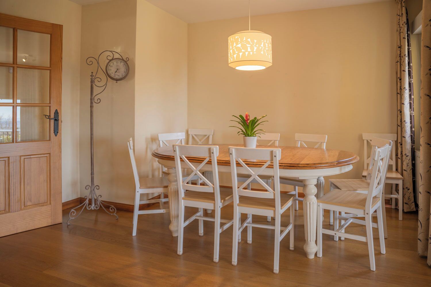 Comedor con mesa de madera y sillas blancas, una lámpara colgante y un reloj en la pared.