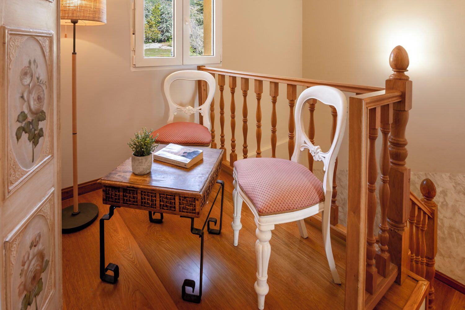 Ein Zimmer mit Holzboden, zwei weiße Stühle mit roten Kissen, ein kleiner Tisch mit einem Buch und einer Pflanze, eine Stehlampe und eine Treppe mit hölzernen Geländern.