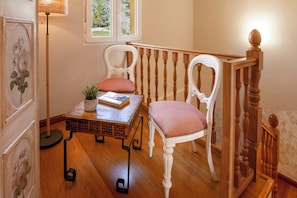 Ein Zimmer mit Holzboden, zwei weiße Stühle mit roten Kissen, ein kleiner Tisch mit einem Buch und einer Pflanze, eine Stehlampe und eine Treppe mit hölzernen Geländern.