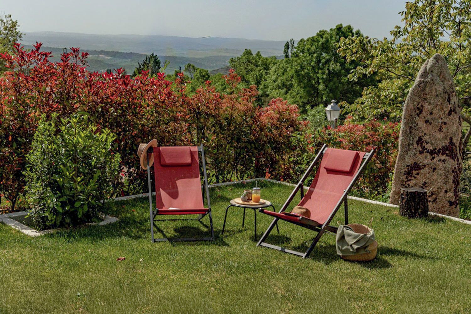 Dos sillones rojos con una mesita y una bebida, sobre un césped con vistas a un paisaje montañoso.