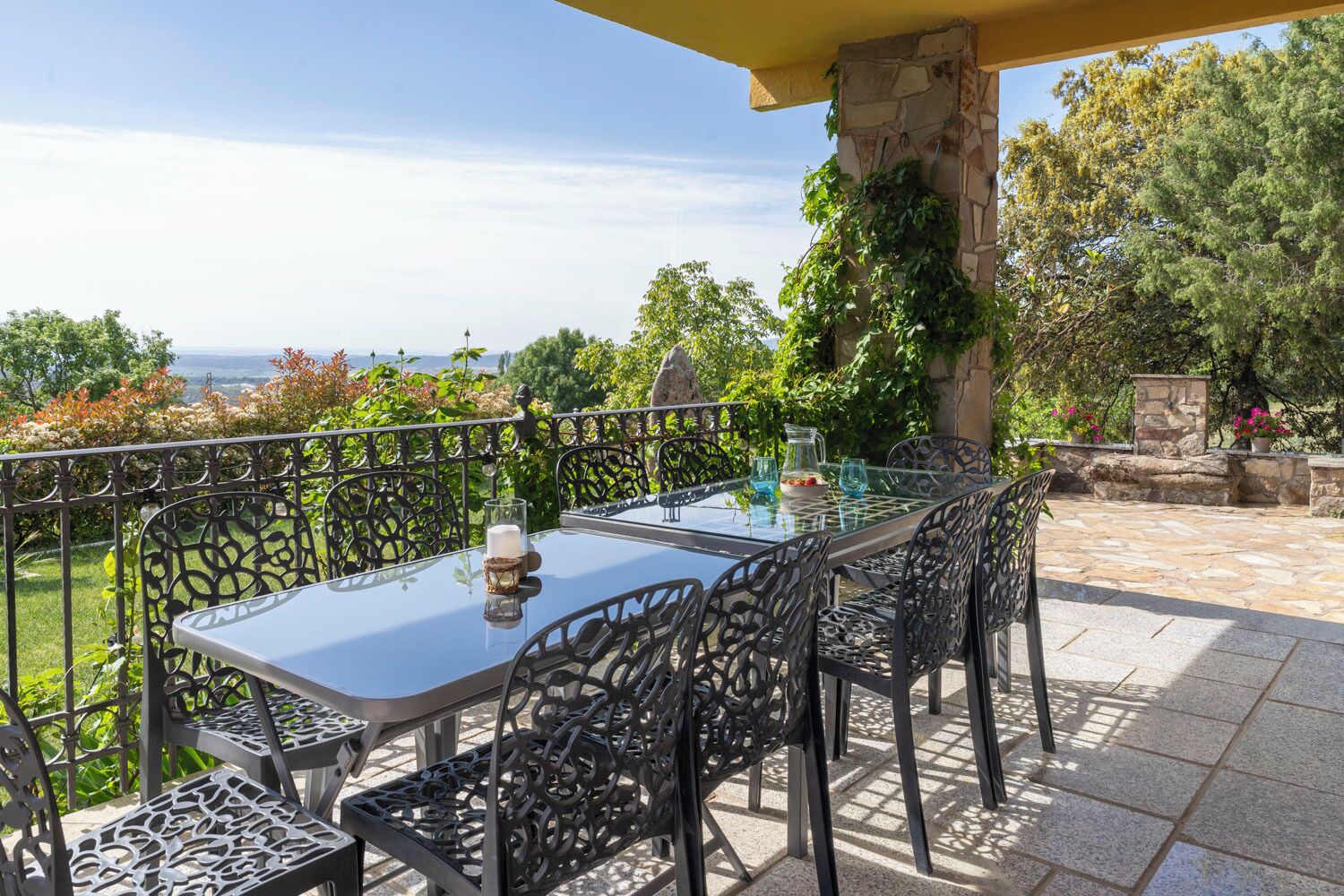 Un patio exterior con una mesa de vidrio y sillas de metal, rodeado de muros de piedra y follaje.