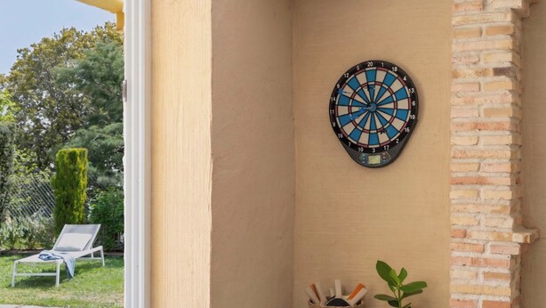 Eine Dartscheibe an einer Wand, darunter ein kleines Regal mit einem Korb voller Darts und einem Blumentopf.