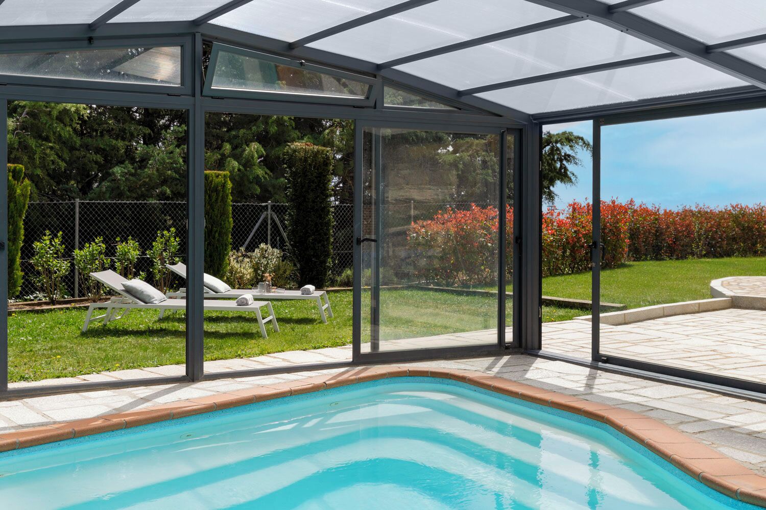 A swimming pool with a glass enclosure, lounge chairs, and a view of a garden.
