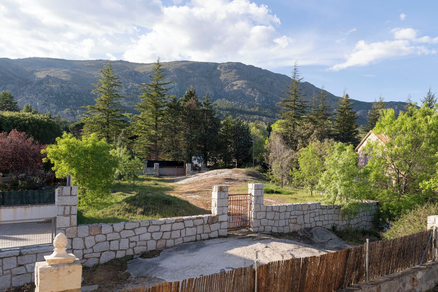 Un muro de piedra con una verja que da a una zona cubierta de hierba, con árboles y una montaña al fondo.