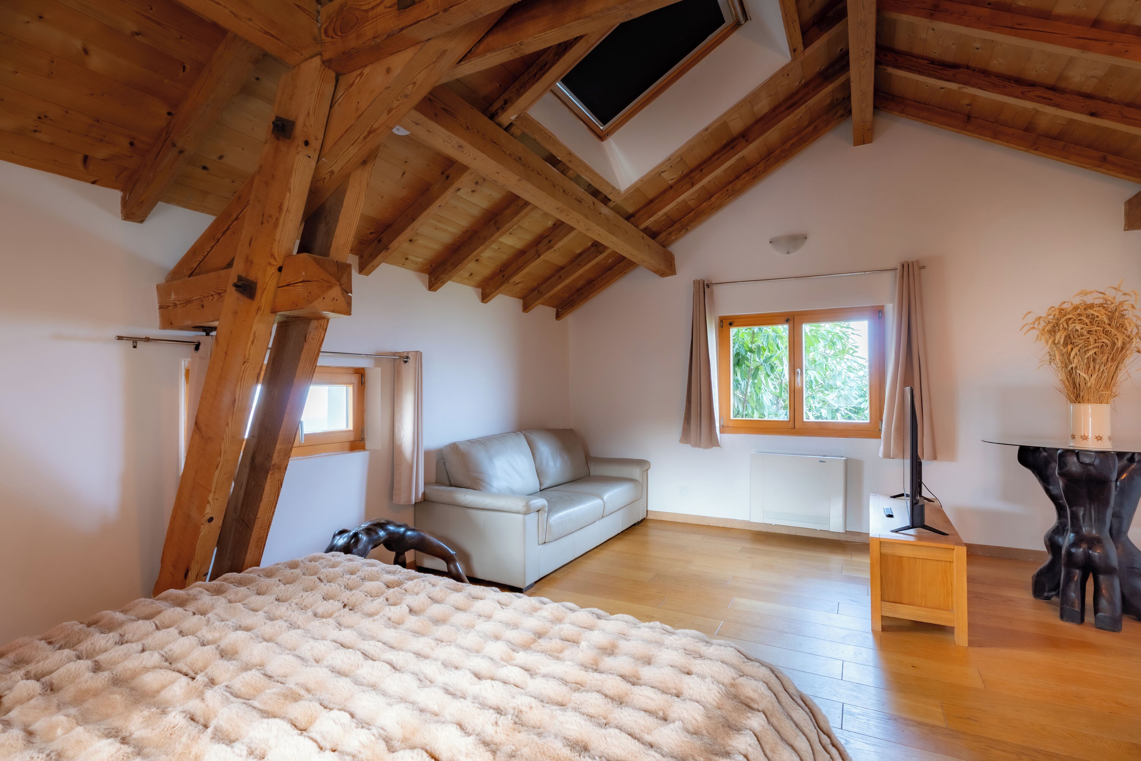 A room with a sloped wooden ceiling, a bed with a fluffy blanket, a leather sofa, a small table with a vase, and a window with curtains.