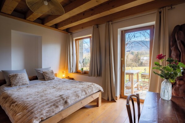 A bedroom with a wooden bed, a bedside table, a window with curtains, and a view of the outdoors. - Vrbo Property (Neydens)