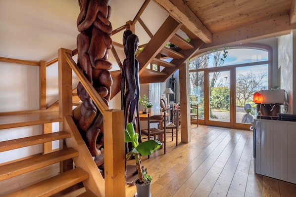 Een houten trap met een uniek, handgesneden detail, een eetgedeelte met tafel en stoelen, en uitzicht door grote ramen.