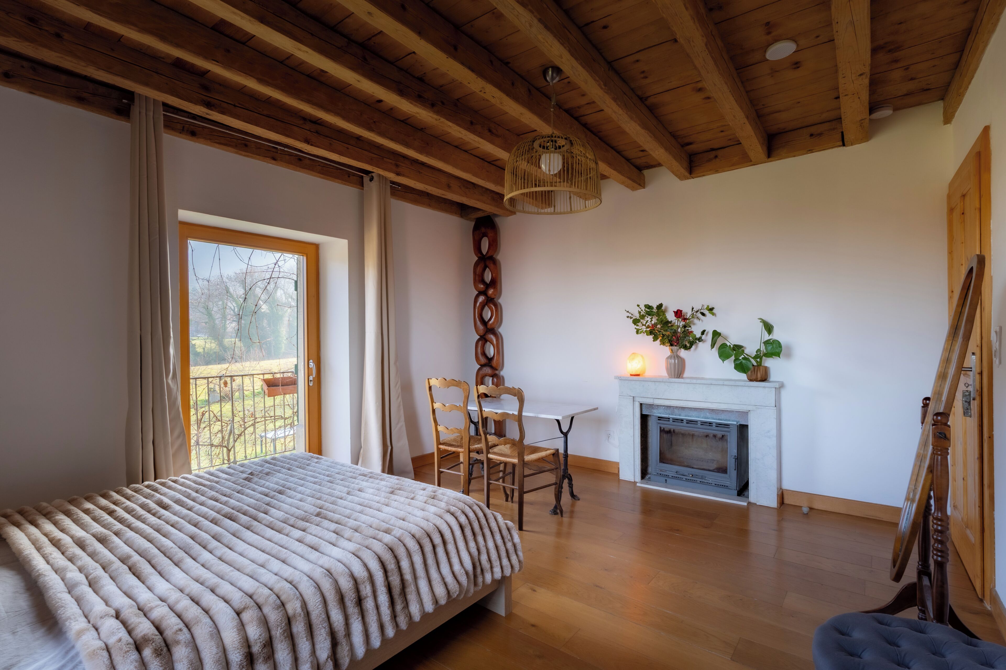 Um quarto com cama, teto de madeira, lareira, mesa com cadeiras e vista pela janela.